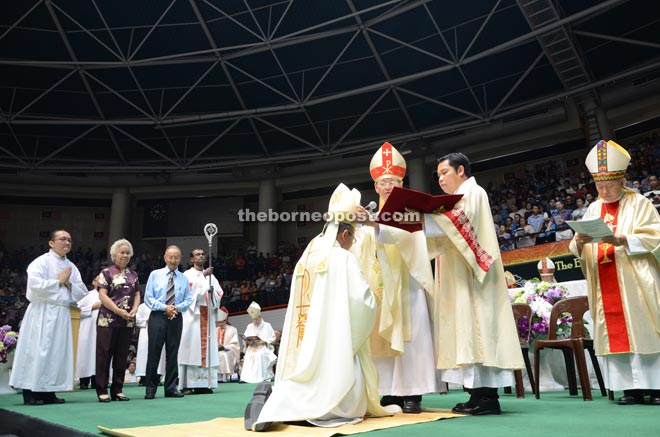 Ha places a Miter (hat worn by bishops) on Poh’s head during the ordination ceremony. On the far left are Poh’s parents witnessing the ordination of their son on stage.