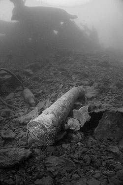A single bombshell sits on the debris-covered seabed, surrounded by mangled metal.