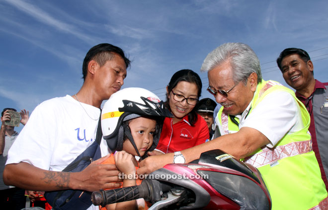 Masing helps a child to put on a helmet when officiating at the state-level Gawai Dayak Road Safety Safety Advocacy 2016 programme. Seen at third left is Davina. — Photo by Chimon Upon 