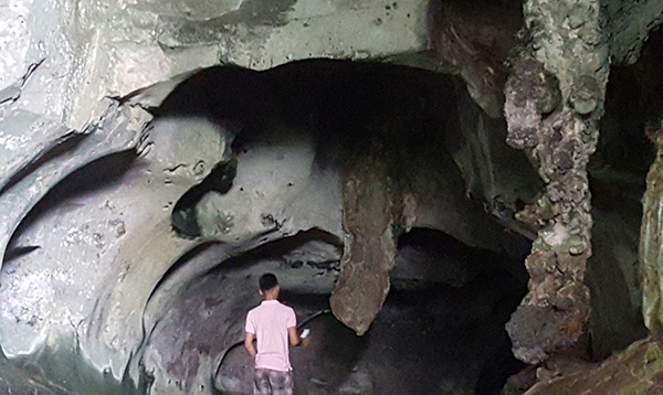 A visitor stands near a mouth-like opening in the cave with a uvula-shaped rock formation jutting down from the roof.