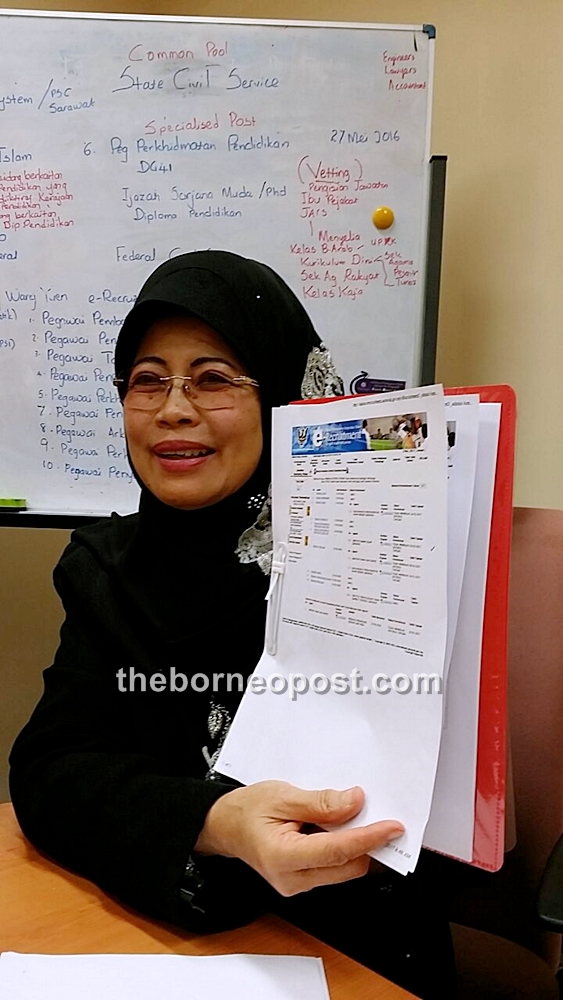 Fatimah holds up a printed image of the e-recruitment website, with a whiteboard explaining e-recruitment and SPP in the background.