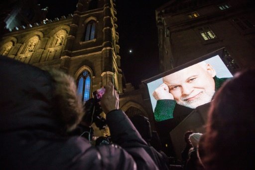 AFP/File / by Marc Braibant | A woman takes a photograph of a large screen showing a photo of the late René Angélil, husband and manager of Canadian singer Céline Dion, as hundreds wait in line outside Montréal's Notre-Dame Basilica January 21, 2016 to pay their respects 